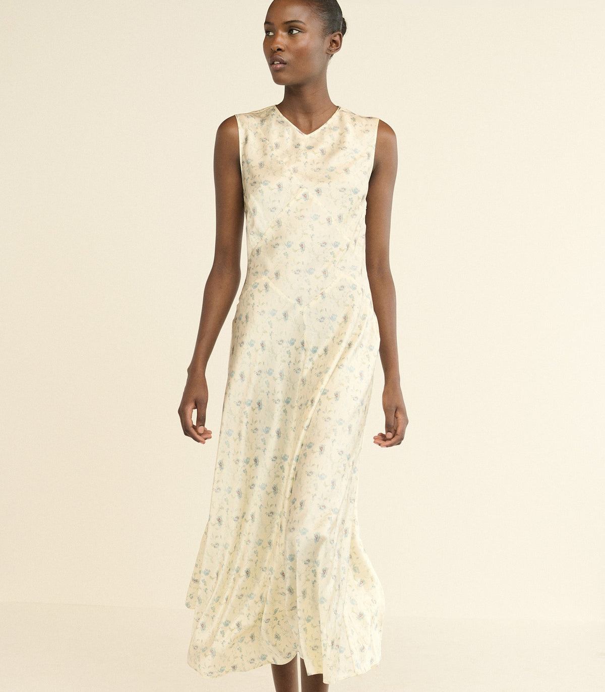 A sleeveless, V-neck midi dress in a cream-colored silk fabric with a delicate floral print, modeled against a solid light beige background.
