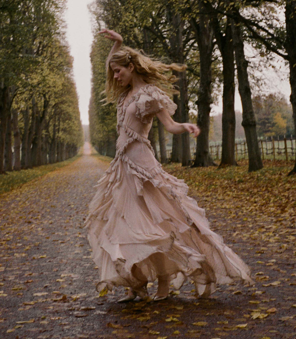 Woman in a long, antique blush dress dancing on a tree-lined path with fallen leaves.