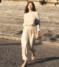 A woman walking outdoors on stone steps wearing a cream-colored two-piece outfit consisting of a sheer long-sleeve crop top with lace detailing and matching loose trousers. She is carrying a small black shoulder bag.