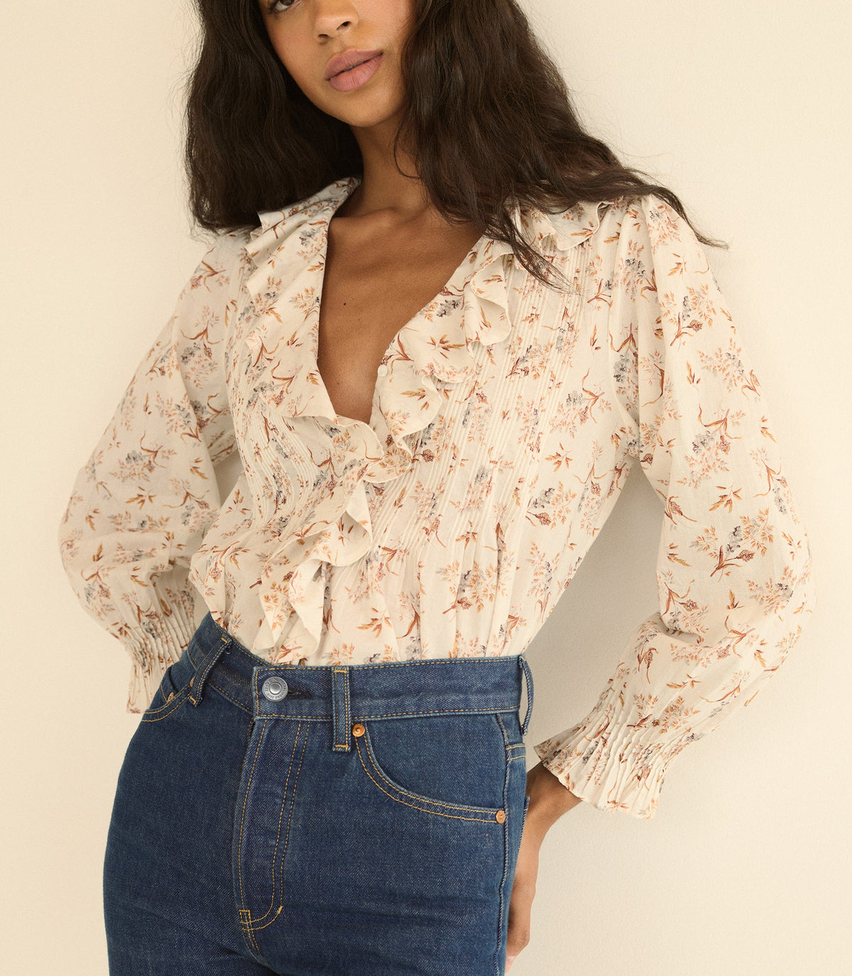 A woman wearing a cream-colored long-sleeve blouse with a delicate orange and blue floral print, paired with high-waisted blue jeans, against a plain beige background.