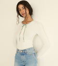 A woman wearing a white ribbed long-sleeve top with a mint blue tie detail at the neckline, paired with light blue jeans, against a plain off-white background.