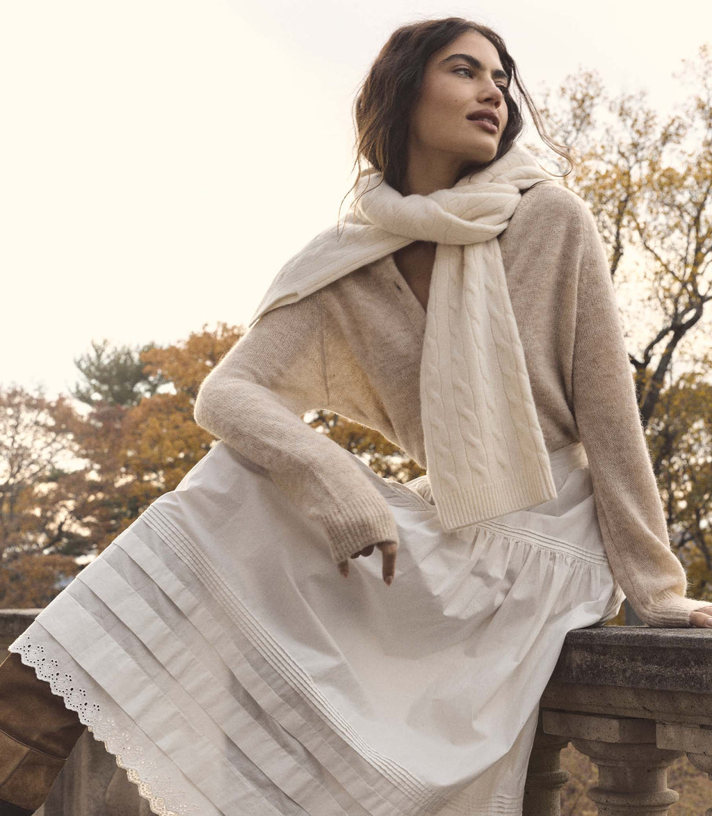Woman wearing a beige sweater and scarf with a white dress, sitting outdoors.