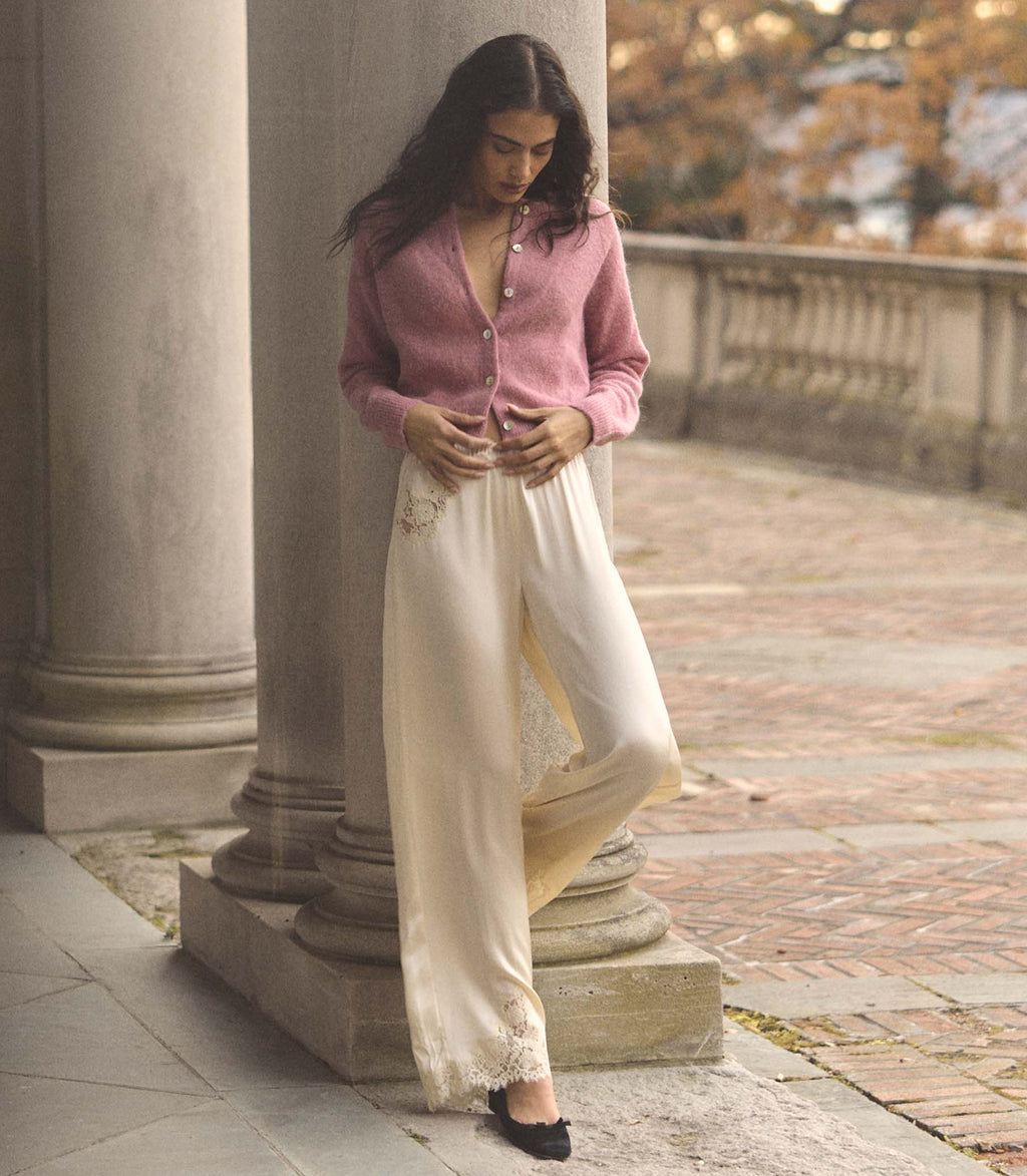 Woman in a pink cardigan and white pants standing on a stone path with columns in the background.