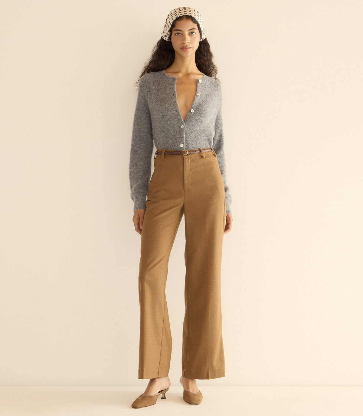 Woman wearing a gray cardigan and brown pants with a beige headband against a plain background