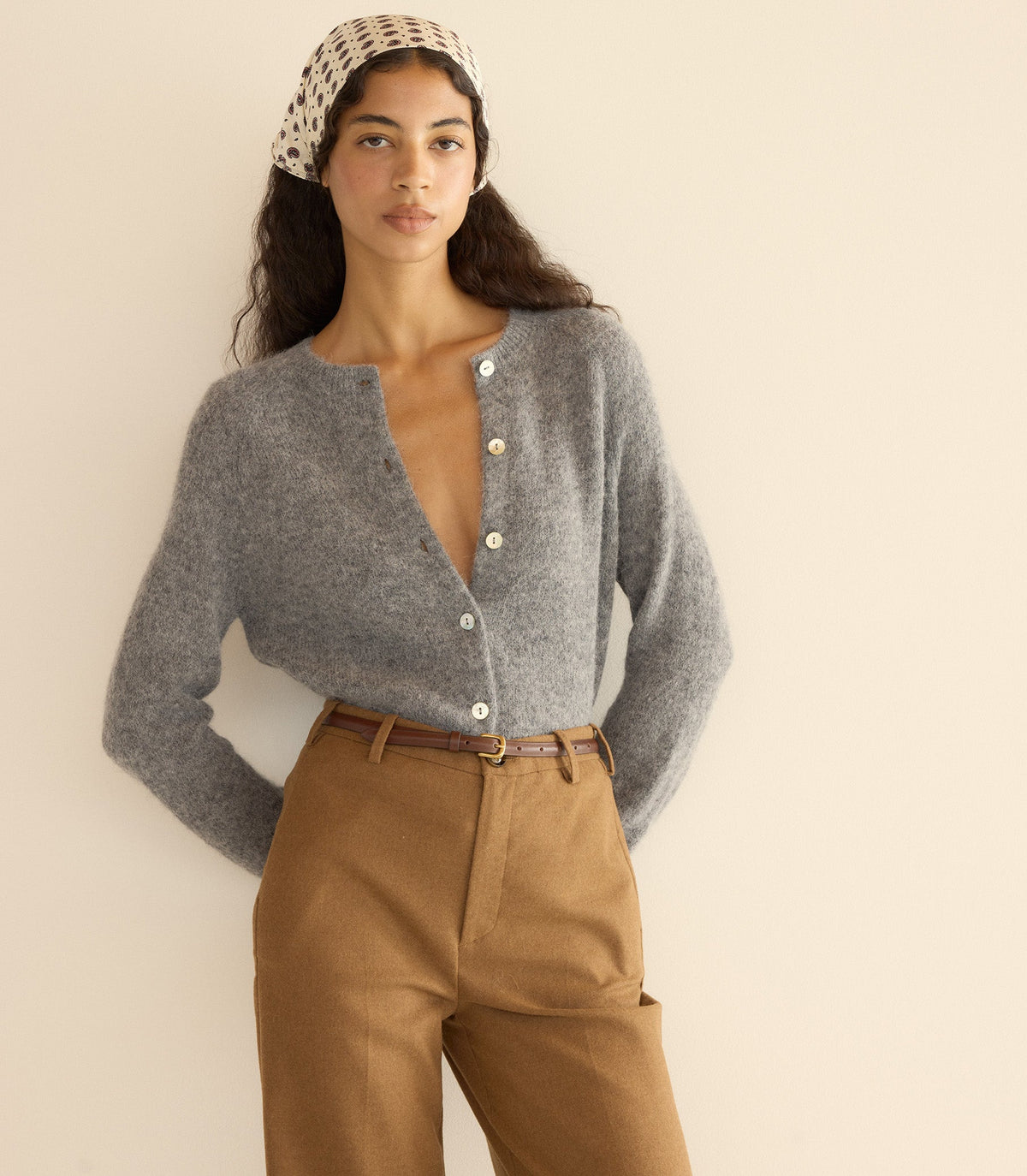 Woman wearing a gray cardigan and brown pants with a beige headband against a plain background