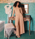 A woman with curly dark hair models a long-sleeved beige lace maxi dress in a vintage-style room with teal mosaic tile walls, leaning on a wooden table.